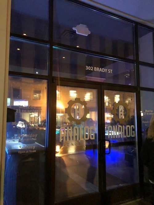 Exterior view of a bar named "Analog" with large glass doors and illuminated signage at night.