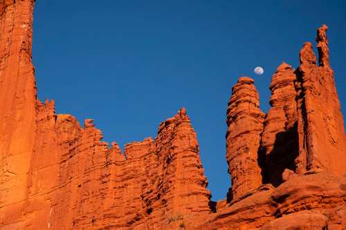 A clear blue sky above towering red rock formations, with a crescent moon visible in the upper right corner.