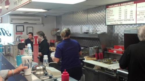 A busy diner counter with staff preparing food, customers ordering, and a menu board displaying prices.