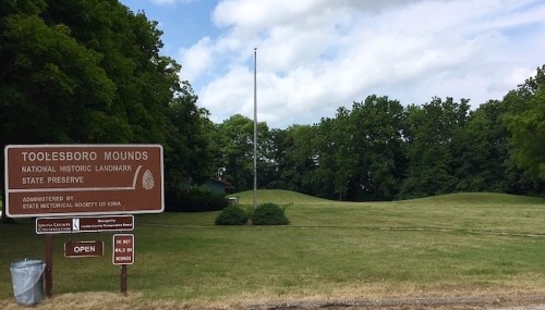 Sign for Toolesboro Mounds, a National Historic Landmark, with grassy mounds and trees in the background under a cloudy sky.