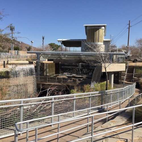 A modern water treatment facility with a flowing stream, surrounded by pathways and trees under a clear blue sky.