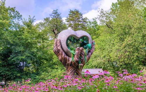 A large heart sculpture held by wooden hands, surrounded by colorful flowers and lush greenery.