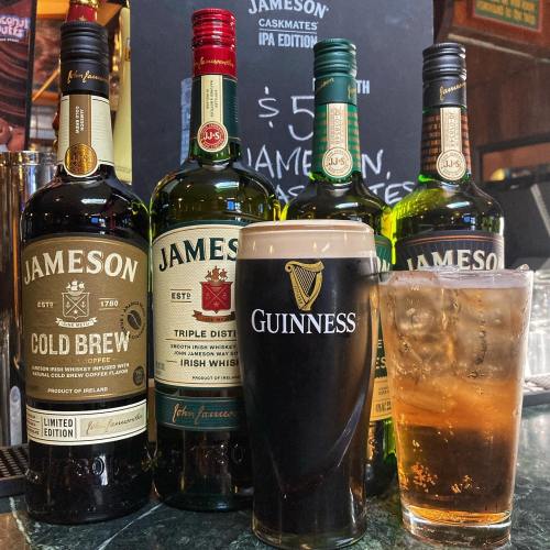 A pint of Guinness and a glass of whiskey sit on a bar, with several bottles of Jameson whiskey in the background.