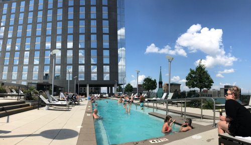 A rooftop pool area with people swimming and lounging, surrounded by tall buildings and a clear blue sky.