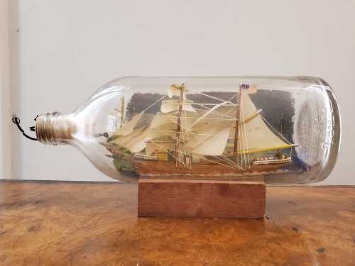 A detailed model ship inside a glass bottle, displayed on a wooden base. The ship features sails and intricate details.