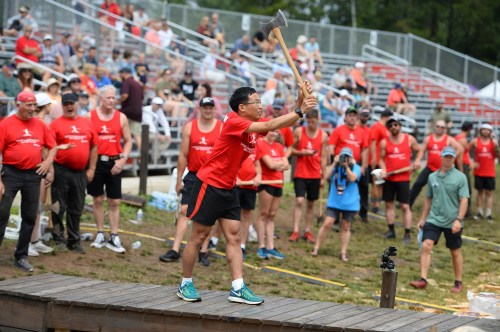 A competitor swings an axe during a lumberjack competition, with a crowd of spectators in red shirts watching.