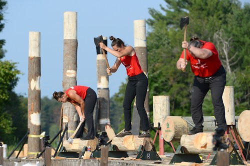 Three women in red shirts compete in a lumberjack event, chopping wood on logs with axes in a forested setting.