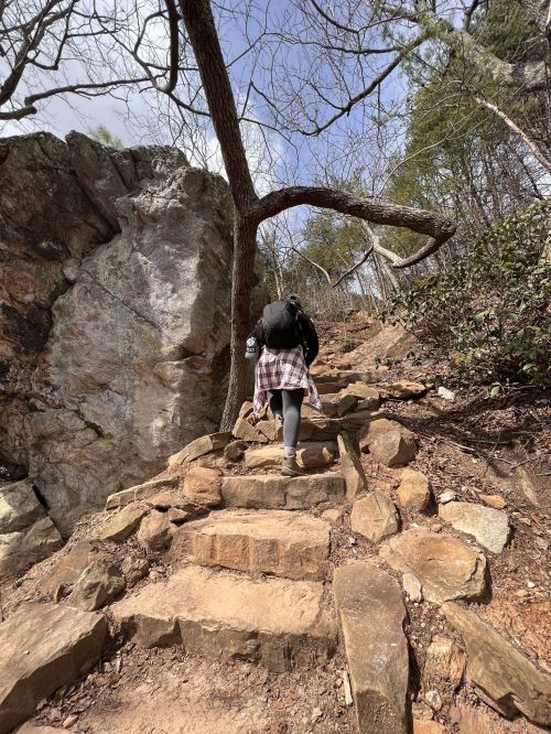 A person walks up a rocky trail surrounded by trees and a large rock formation.