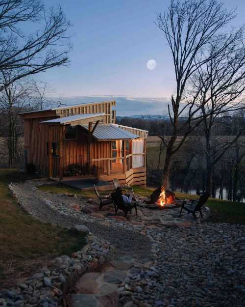 A cozy wooden cabin by a fire pit, surrounded by trees, with a full moon in the evening sky.