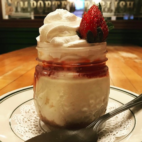 A dessert in a jar with whipped cream, a strawberry on top, and a layer of strawberry sauce, served on a plate.