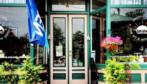 A charming café entrance with a blue flag, potted plants, and large windows displaying art and baked goods.