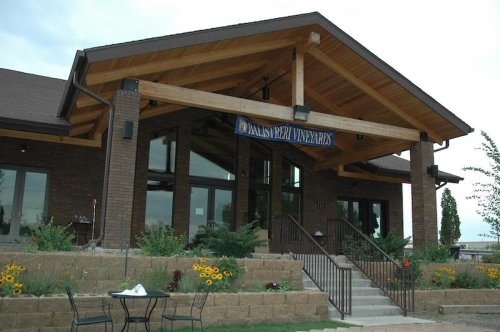 A modern winery building with a wooden overhang, large windows, and a landscaped entrance featuring flowers and seating.