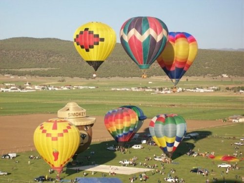 A colorful array of hot air balloons floats above a green landscape, with spectators and vehicles below.