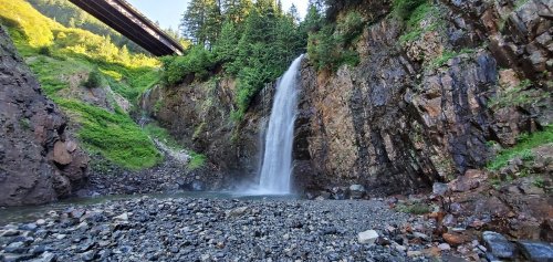A waterfall cascades down rocky cliffs surrounded by lush greenery, with a bridge visible above.