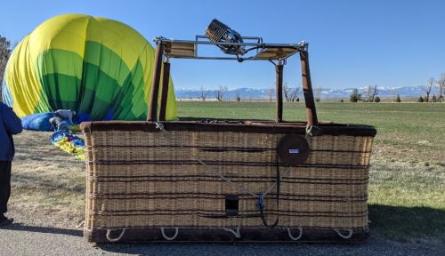 A colorful hot air balloon is partially inflated beside a large wicker basket on a grassy field with mountains in the background.