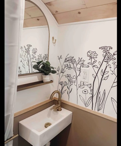 A small bathroom featuring a round mirror, floral wall art, a sink, and a plant on a shelf.