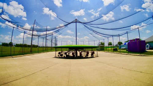 A wide view of a sports facility with a green canopy, surrounded by nets and a clear blue sky with fluffy clouds.