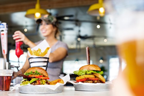 Two burgers with toppings and fries in a basket, with a smiling server in the background pouring ketchup.