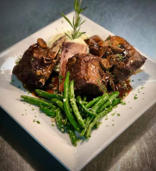 A plated dish featuring tender meat, sautéed mushrooms, green beans, and a garnish of rosemary on a white square plate.