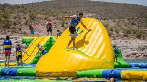 A group of people in life vests play on colorful inflatable water structures in a lake.