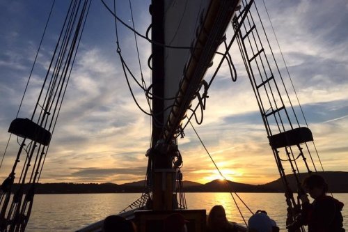 Silhouette of a sailboat against a sunset, with masts and rigging framing the colorful sky and calm water.