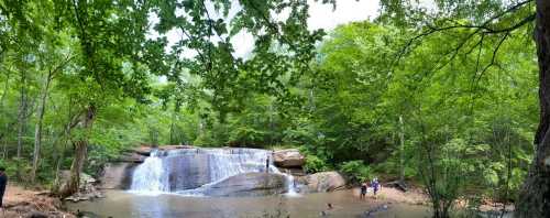 A serene waterfall cascades over rocks, surrounded by lush green trees and people enjoying the water below.