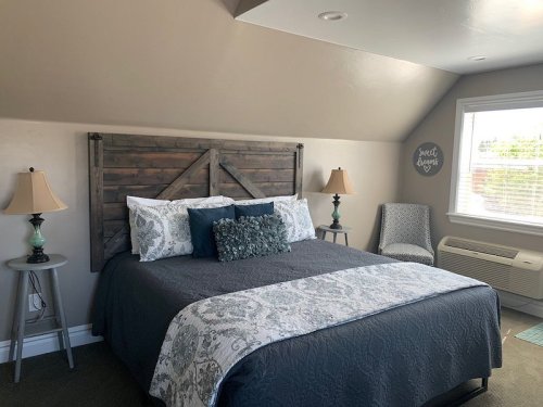 Cozy bedroom with a wooden headboard, blue and gray bedding, two lamps, and a chair by the window.