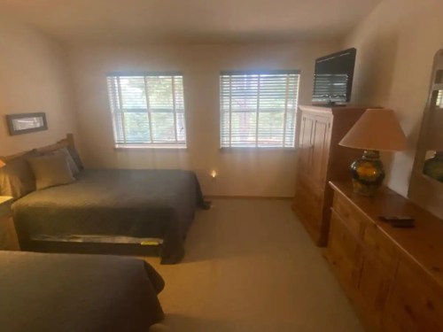 Cozy bedroom with two beds, a dresser, lamp, and windows with blinds, creating a warm and inviting atmosphere.