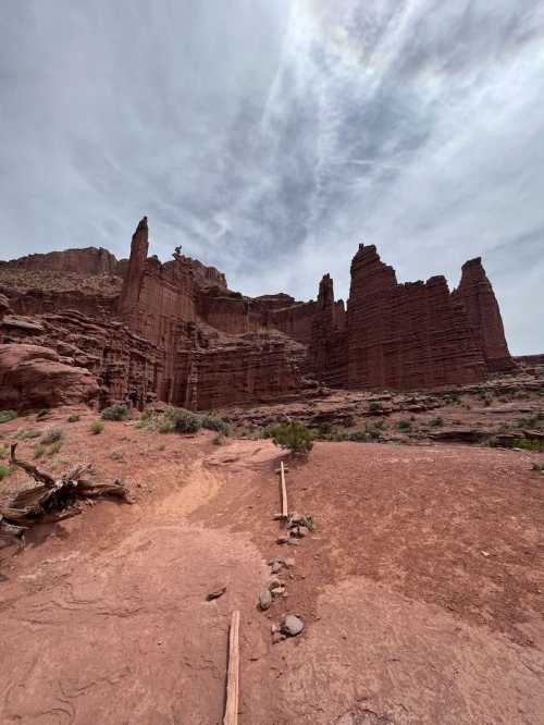 A rocky landscape with towering red cliffs under a cloudy sky, featuring a dirt path and sparse vegetation.