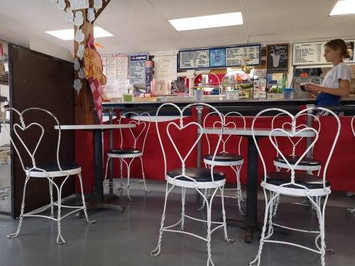 A cozy café interior with heart-shaped chairs, a counter displaying menu items, and a waitress taking an order.