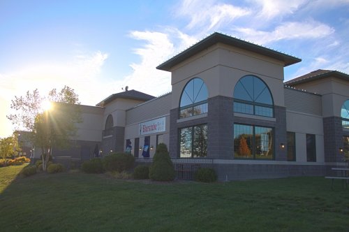 A modern building with large windows, surrounded by greenery, under a clear sky with the sun setting in the background.