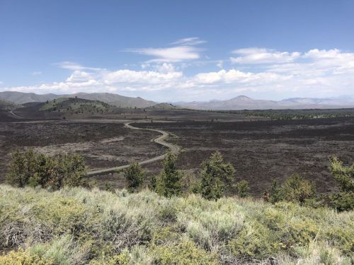 A winding dirt road cuts through a vast, open landscape with sparse vegetation and distant mountains under a blue sky.