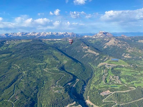 A hot air balloon floats over lush green valleys and mountains under a clear blue sky.