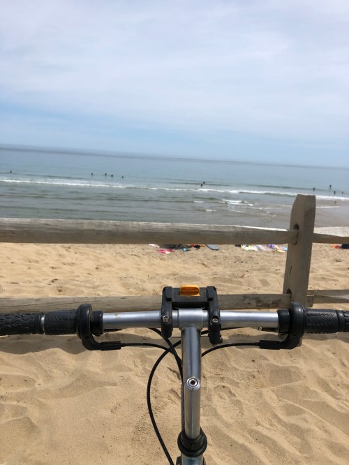 View from a bike handlebar on the beach, with surfers in the ocean and a sandy shore in the background.
