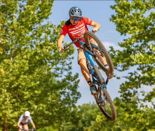 A cyclist performs a jump on a mountain bike, surrounded by trees and a clear blue sky.