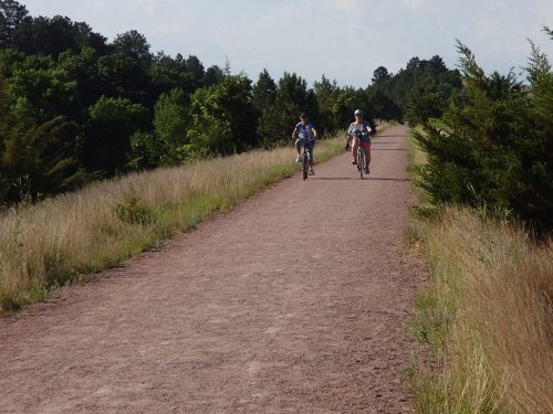 Two cyclists ride along a dirt path surrounded by greenery and trees on a sunny day.