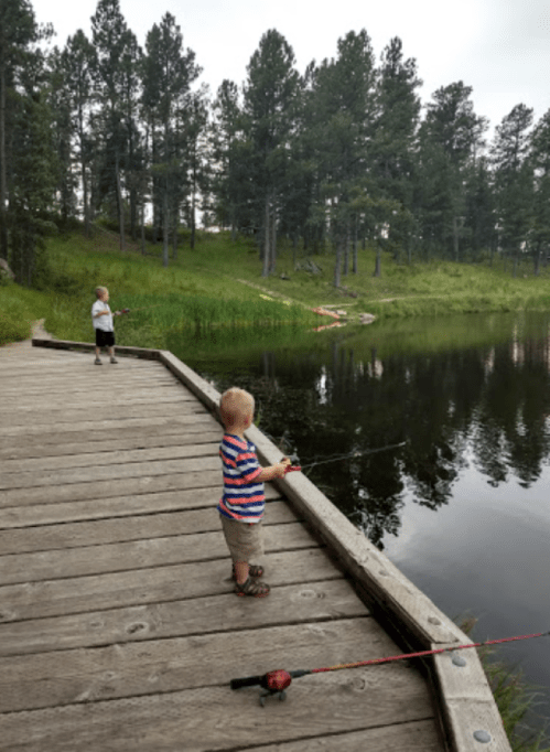 Two young children fishing from a wooden dock by a calm lake, surrounded by trees and greenery.