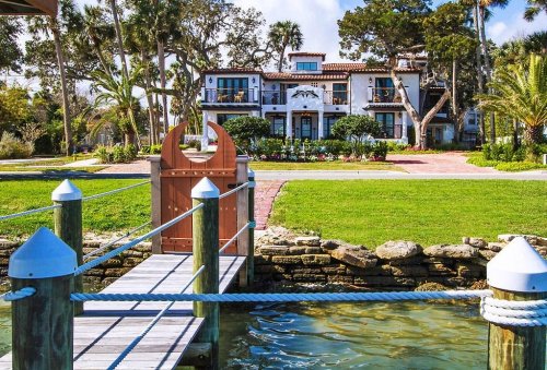 A waterfront view of a large, elegant house surrounded by lush greenery and palm trees, with a wooden dock in the foreground.