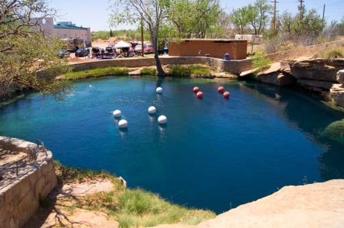 A clear blue pond surrounded by rocks and trees, with floating buoys in the water and a gathering area in the background.