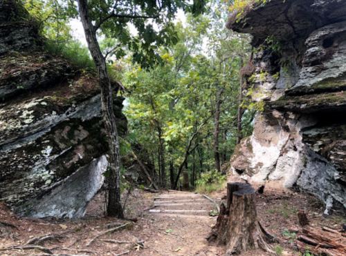 A narrow trail between large rock formations, surrounded by trees and greenery.