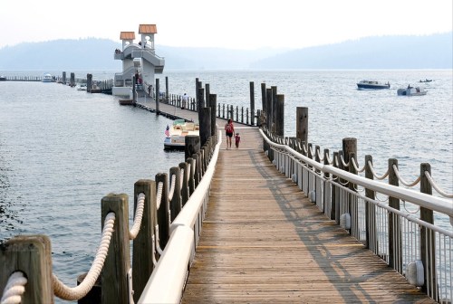 A wooden pier extends into a calm lake, with people walking and boats in the distance under a hazy sky.