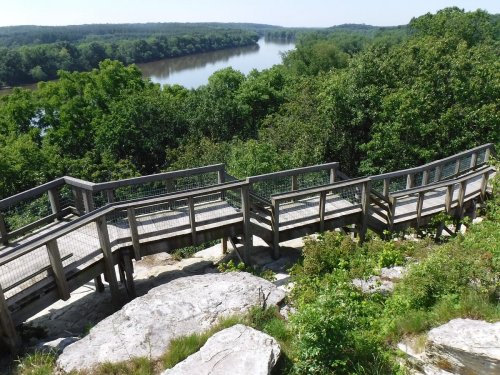 A winding wooden walkway leads down to a river, surrounded by lush green trees and rocky terrain.