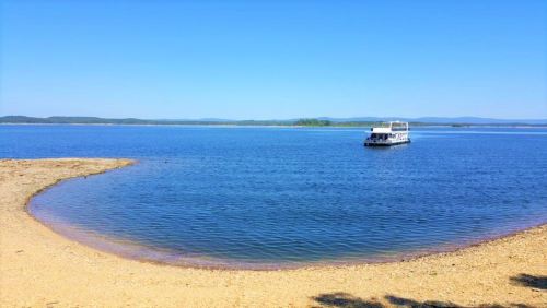 A serene lake scene with a sandy shore and a boat floating on calm blue waters under a clear sky.
