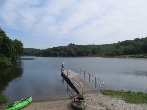 A calm lake scene with a person fishing from a dock, surrounded by trees and two kayaks on the shore.