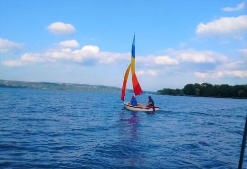 Two people sail on a colorful boat on a calm lake under a blue sky with fluffy clouds.