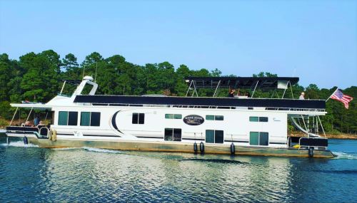 A large houseboat cruising on calm water, surrounded by green trees, with people enjoying on the upper deck.