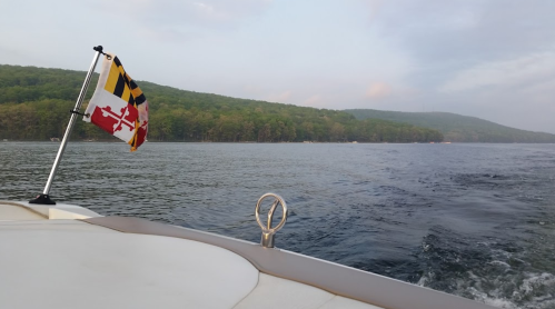 A boat's view of calm water with a Maryland flag flying, surrounded by green hills under a cloudy sky.