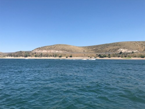 A serene view of a blue lake with gentle waves, surrounded by rolling hills under a clear blue sky.