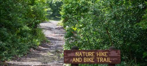A wooden sign reading "Nature Hike and Bike Trail" at the entrance of a lush, green path surrounded by foliage.