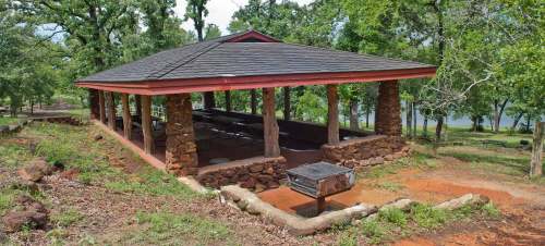 A stone pavilion with a red roof surrounded by trees, featuring a grill area and a view of a nearby lake.